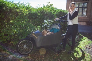 Frau steht mit Cargobike vor einer Hecke, im Fahrrad sitzt ein Hund